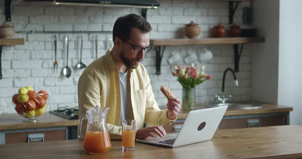Young Man Freelancer in Eyeglasses Working at Laptop Computer and Eating Tasty Croissant with Juice