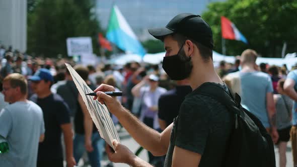 Male Protester Drawing Banner Sign in Hands with Marker in Rally Riot Crowd