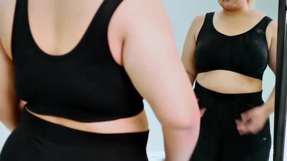 Overweight young woman in sportswear looking her belly fat into the mirror