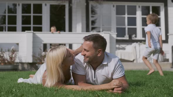 Loving Mom and Dad Kiss Lying on the Grass Near the House While the Children Run Around Them