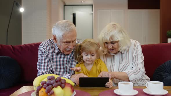 Grandfather and Grandmother Sitting in Living Room and Teaching Small Granddaughter Using Tablet