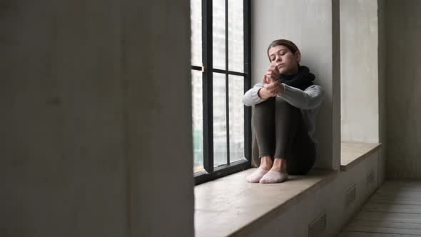 A sickly-looking young woman is sad on the windowsill of a city apartment