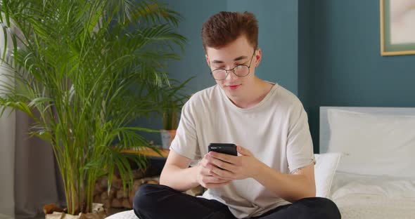 Young single man using dating app on his phone, swiping and touching screen, at home on bed.