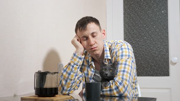 Sleepy Man Sits with Gray Cat at the Table in the Kitchen