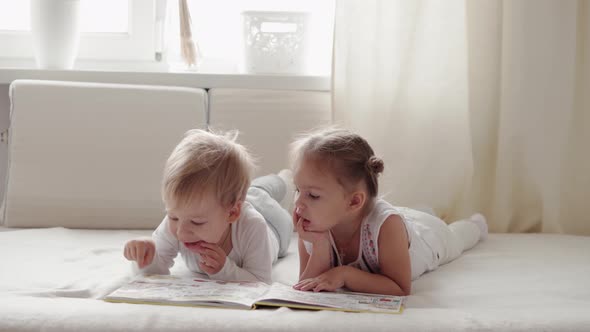 Two smiling children read large interesting book of fairy tales on bed.