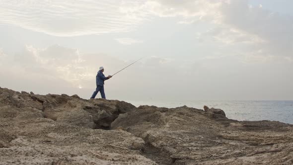 Old fisherman standing on sea side rocks and fishing against the sunset