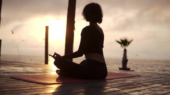 Yogi Girl is Sitting in Lotus Pose in Front the Sea in Summer on Sunset and Meditating Seagulls