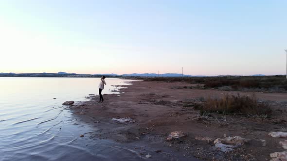 Man Hipster with Guitar Runs Towards the Waterfront and Jumps Up Raising Arms Up  Aerial