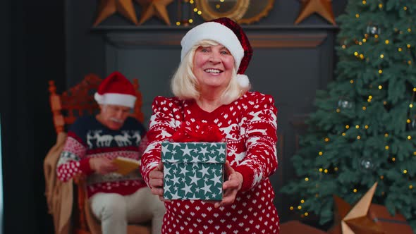 Senior Grandmother in New Year Sweater and Hat Presenting Christmas Gift Box Holidays at Home