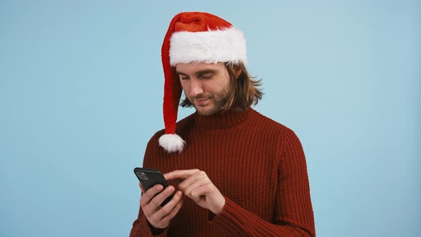 Young Male in Santa Hat is Holding Smartphone and Using It Smiling and Showing Thumb Up Posing on