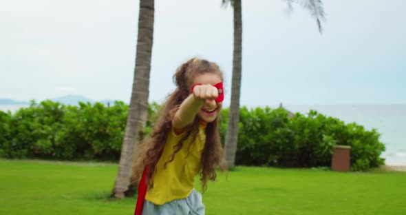 Portrait of a Cute Funny Child a Little Girl in a Superhero Costume in a Red Cloak and a Red Mask