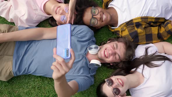 Top View Portrait of Smiling Cheerful Multiethnic Friends Lying Together on Lawn Taking Selfie