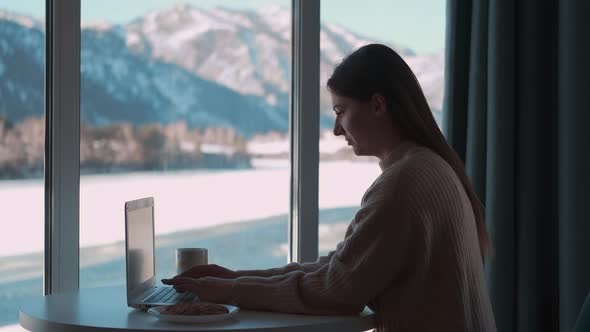 Beautiful Female Freelancer Working From Home Using a Laptop While Sitting at a Table By the Window