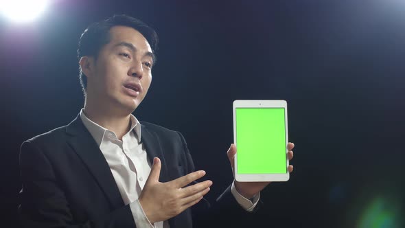 Asian Speaker Man In Business Suit Holding And Pointing Green Screen Tablet While Speaking