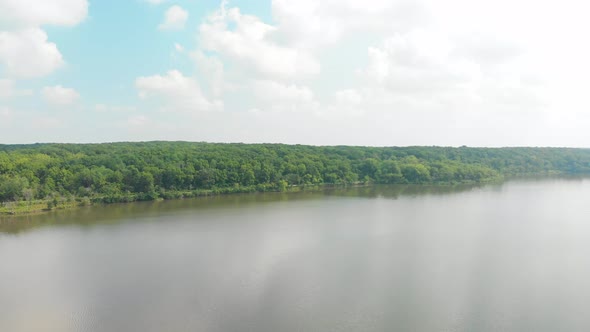 drone forward flight to a horizon and a tree line over a lake with bright green leaves with white pu