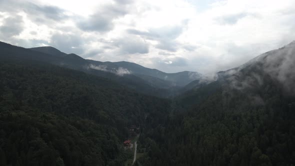 Flying into valley covered in pine forest and low hanging clouds with lone house standing at the end