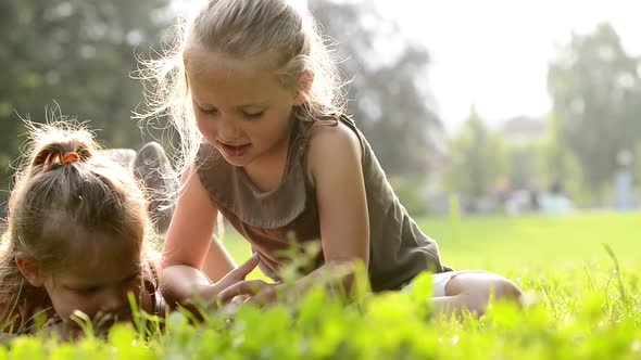 Happy Blonde Sisters Lay Down on Grass are Reading Fairy Tales on Tablet at Town Park in Summer Day