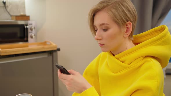 a Young Woman in a Yellow Sweatshirt is Sitting in the Kitchen with a Serious Face Looking at