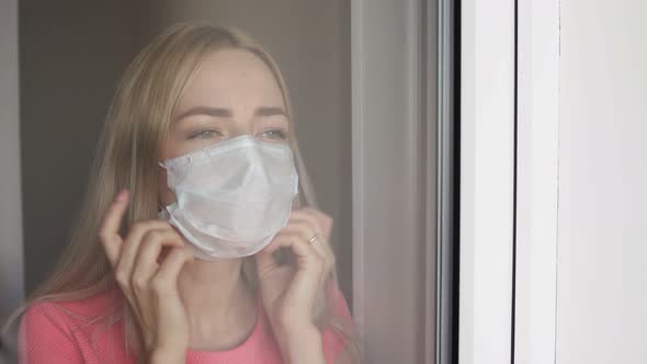 Home Quarantine. Caucasian Woman Standing at Window in a Medical Mask, Looking Out.
