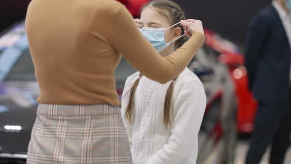 Portrait of Cute Brunette Girl with Unrecognizable Slim Woman Putting Face Mask on Daughter in Car