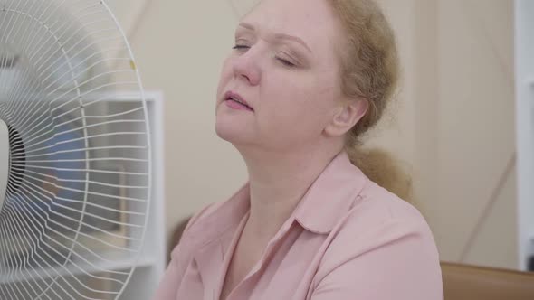 Close-up of Senior Woman Using Electric Fan Indoors. Portrait of Ill Caucasian Lady Having Fever or