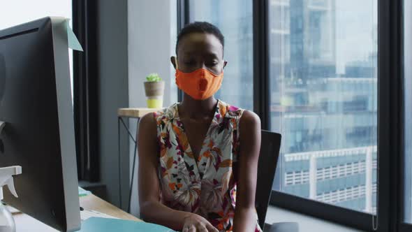 African american woman wearing face mask reading file of documents while sitting on her desk at mode
