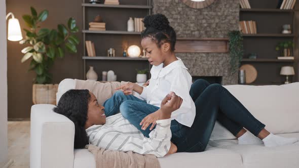 Happy Relaxing African Mother Lying on Sofa and Small Child Active Playful Daughter Girl on Couch