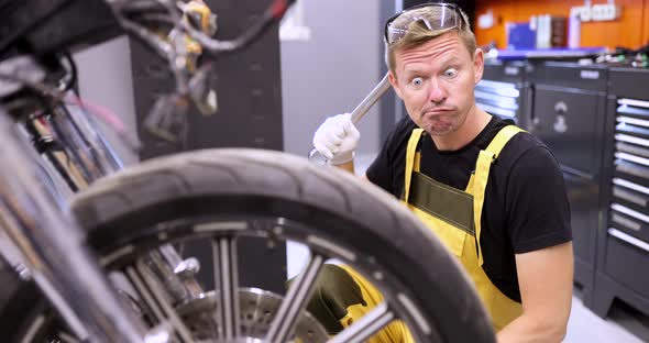 Emotionally Pensive Auto Mechanic Holds Wrench and Stares in Surprise at Motorcycle in Service