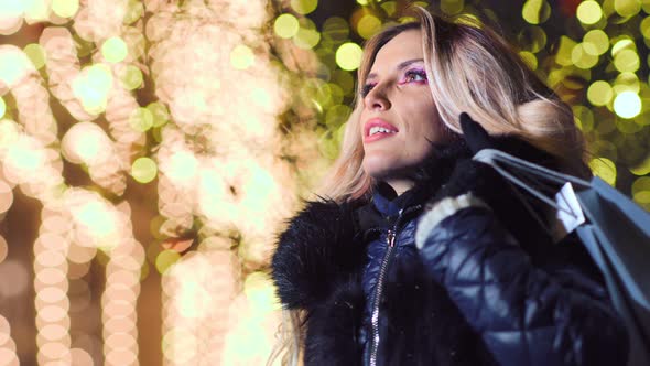 Amazing Beautiful Woman with Christmas Shopping Bags Gifts at Light Bokeh Illuminating Background