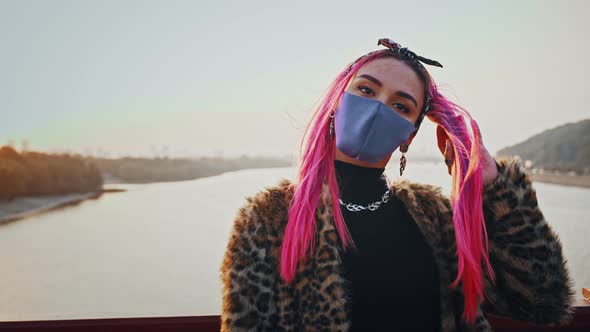 Young Hipster Lady in Informal Clothes and Protective Mask is Standing on Bridge Touching Her Pink