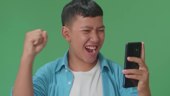 Young Asian Boy Celebrating With Mobile Phone While Standing On Green Screen In The Studio