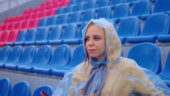 Young woman in raincoat sitting on stadium bleachers alone in rainy weather. 