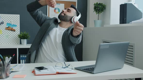 Funny Businessman Wearing Headphones Dancing and Working with Laptop Sitting at Desk in Office