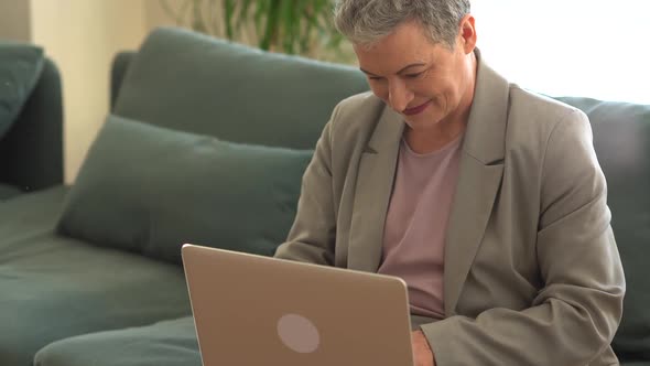 Busy Grayhaired Lady in a Business Suit is Working with a Laptop at Home Alone While Sitting on the