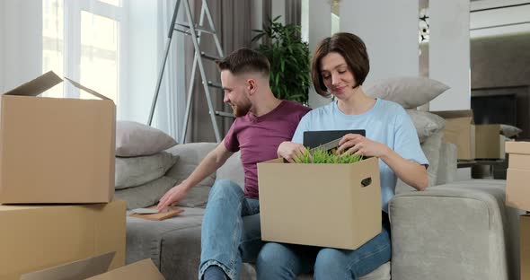 Family Unpacking Cardboard Box with Pictures in Frames Sit on Sofa in New Apartment on Moving Day