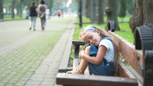 Small tired child girl sitting on a bench with closed eyes resting in summer park.