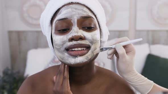 African American Woman with Towel on the Head while Cosmetologist Hand Applying Mask on Her Face