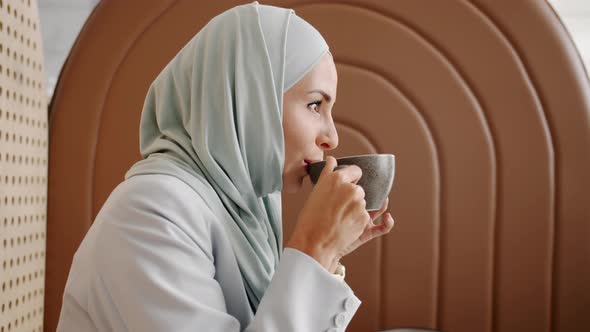 Side View of Beautiful Young Woman Wearing Hijab Drinking Coffee and Smiling in Cozy Cafe