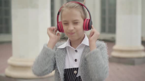 Portrait of Positive Caucasian Schoolgirl Listening To Music in Headphones Standing Outdoors. Happy