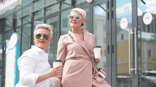 Two Businesswomen Standing Outside with Coffee with Buildings on Background