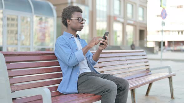 Young African Man Browsing Internet on Smartphone While Sitting Outdoor on Bench