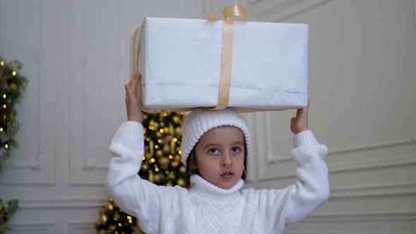 Child Boy in a White Knitted Sweater and Hat Holds Gift Box with a Gold Ribbon on His Head