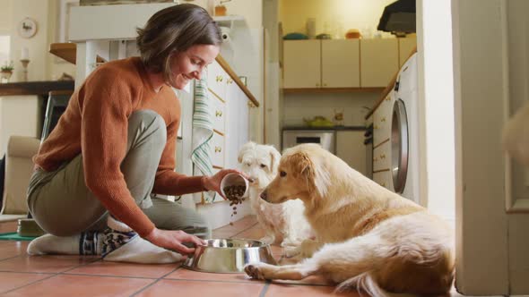 Smiling caucasian woman feeding her pet dogs pouring food into bowl in kitchen at home