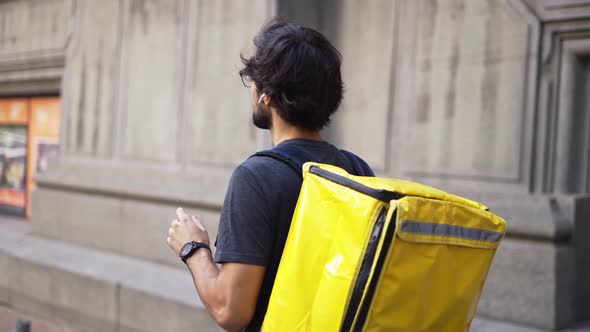 Delivery Man with Yellow Backpack Talk to a Client on the Street Using Earphones Side View