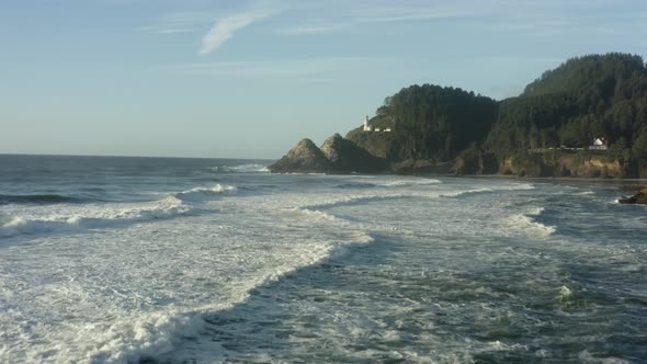 Wide aerial moving low across waves toward Haceta Head lighthouse in Oregon