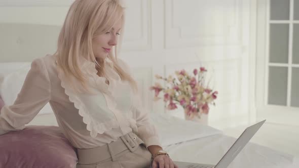 Smiling Blond Caucasian Businesswoman Typing on the Laptop Sitting on the Bed at Home