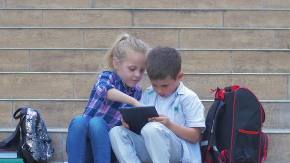 Schoolchildren Use Digital Tablet Sitting on Steps with Backpacks During Recess in Open Air