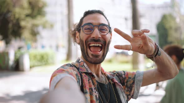 Cheerful curly-haired bearded man in eyeglasses showing peace sign at the camera