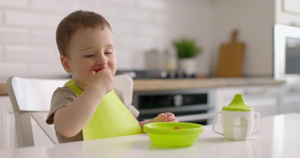 Cute Little Baby Boy Sits at a Table in the Kitchen and Eats Strawberries From Green Plate with His