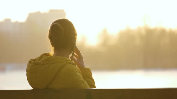 Young Pretty Woman Sitting on a Park Bench Talking on Her Smartphone Outdoors in Warm Autumn Evening